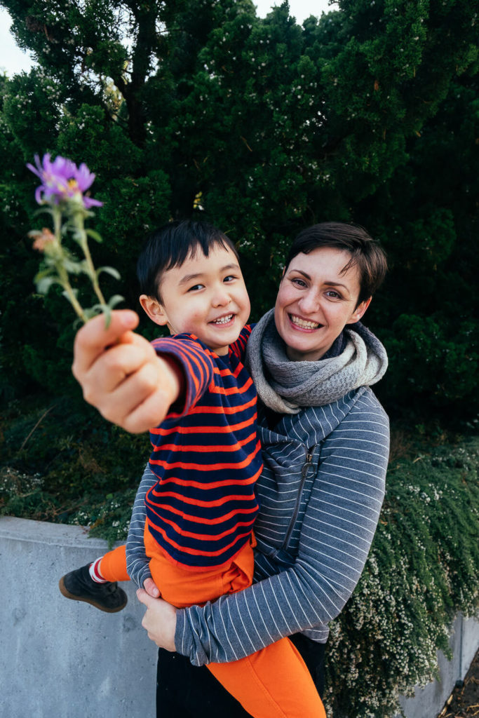 A smiling woman holds a young boy outdoors. The boy, in a red and blue striped shirt and orange pants, offers a purple flower to the camera. Captured by a Brooklyn family photographer, they stand amidst lush green foliage, embodying the warmth of family photography NYC.