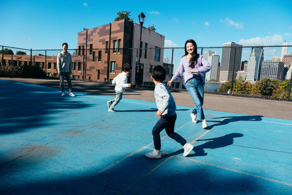 A group of four people enjoying a sunny day on a blue basketball court captures the essence of family photography in NYC. Two children playfully run while two adults watch them. In the background, trees, a building, and a glimpse of a city skyline shimmer under the bright blue sky.