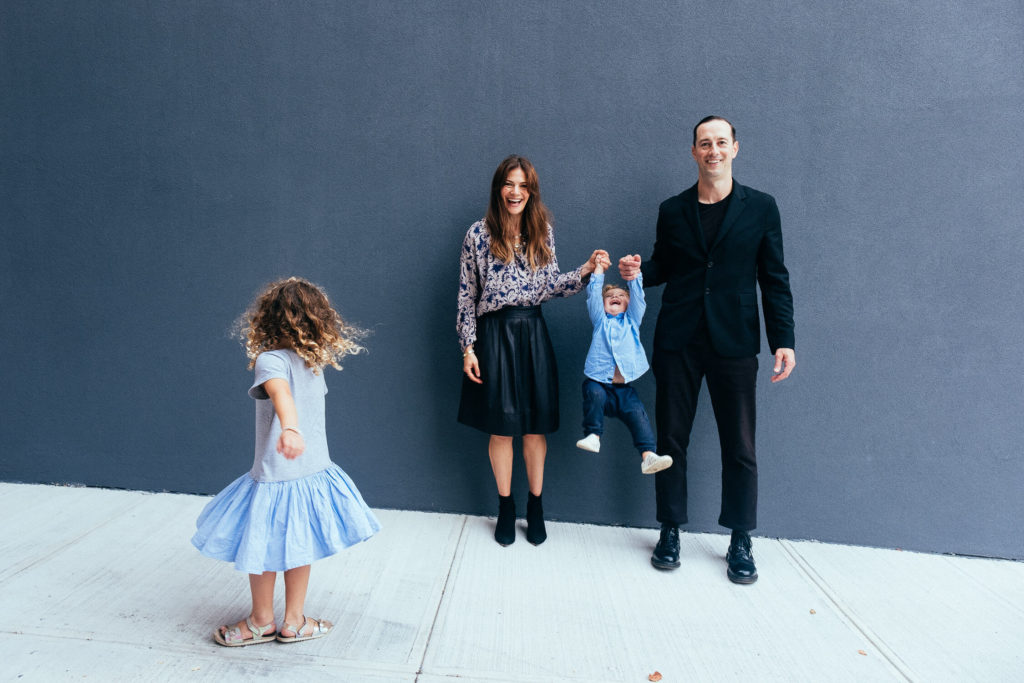 A family stands in front of a gray wall, captured beautifully by a Brooklyn family photographer. A woman and man hold up a cheerful baby between them, while a young girl in a blue dress twirls joyfully. The woman wears a patterned blouse with a black skirt, and the man is dressed in black.