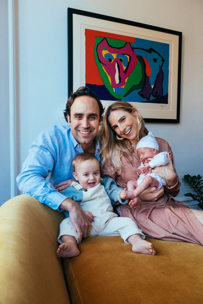 A smiling family sits on a yellow couch, captured by a talented Brooklyn family photographer. The man in a blue shirt and the woman in a pink dress hold two young children—a toddler and a baby. A colorful abstract painting hangs on the wall behind them, adding charm to this moment of family photography in NYC.