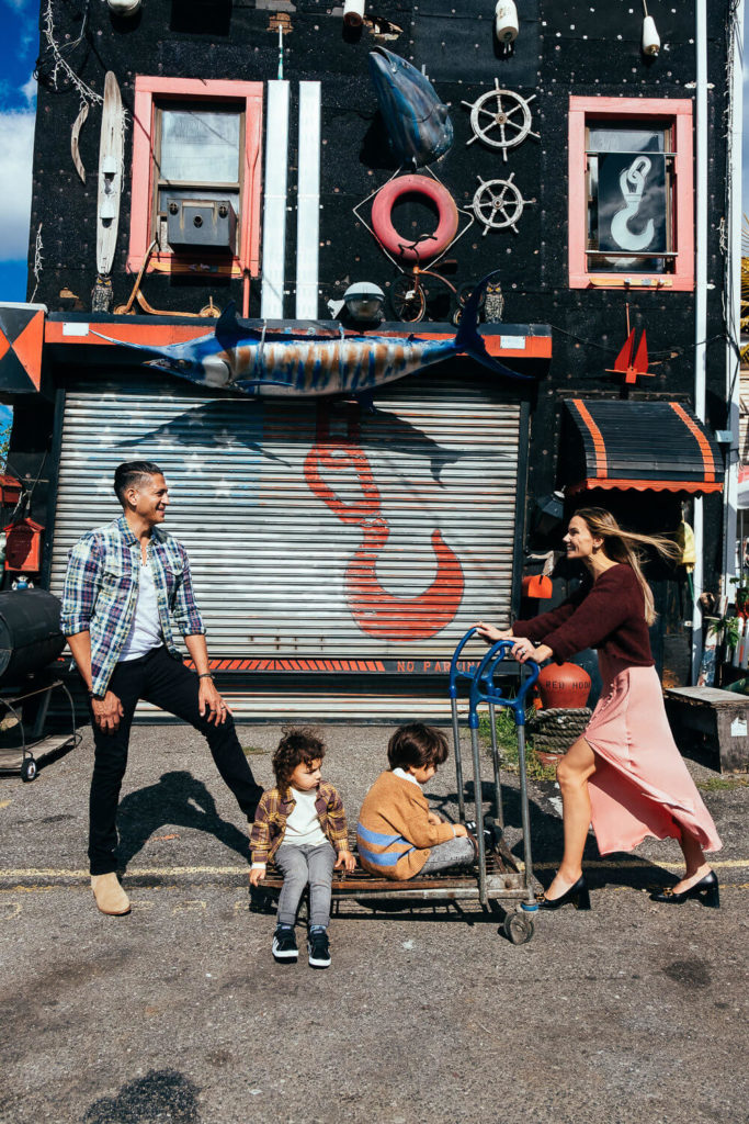 A woman in a pink skirt pushes a cart with two children seated on it as a man stands nearby laughing. The vibrant background, captured by a Brooklyn family photographer, features nautical decorations like a large fish and life preserver. A perfect moment of family photography in NYC.