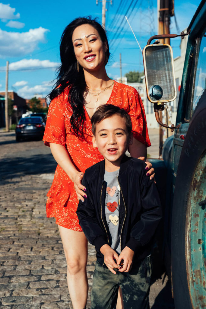 A woman in a red dress stands with a young boy in a black jacket, their smiles captured perfectly by a Brooklyn family photographer. They're posed next to a vintage vehicle on a sunny cobblestone street, the blue sky dotted with clouds—an enchanting moment of family photography NYC.