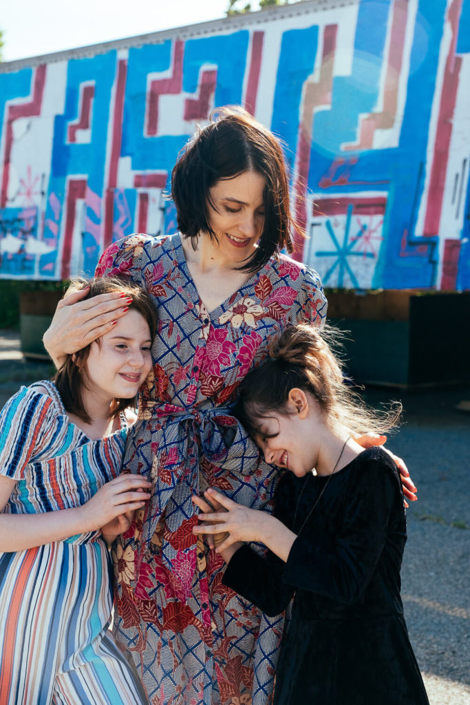 A woman in a floral dress stands with two young girls in striped and black dresses, all smiling for their Brooklyn family photographer. They are posed in front of a colorful, graffiti-covered wall that perfectly captures the vibrant essence of family photography NYC.