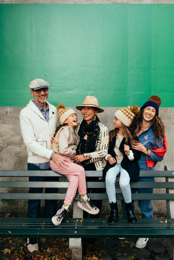 A family of five sits on a park bench, dressed in fall clothing with hats and jackets. Captured by a Brooklyn family photographer, the three adults and two children smile joyfully against a green and gray wall, with leaves beautifully scattered on the ground.