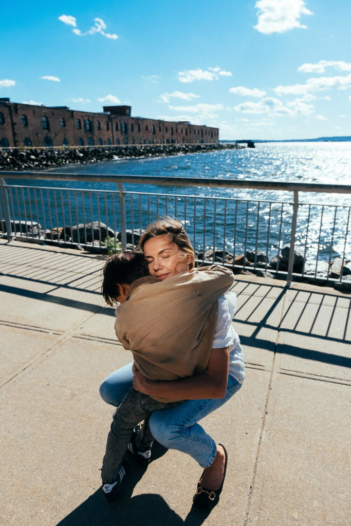 A woman kneels on a sunny waterfront sidewalk, embracing a child in a brown jacket. Captured by a Brooklyn family photographer, the serene scene showcases a large building and calm waters in the background, while shadows grace the concrete.