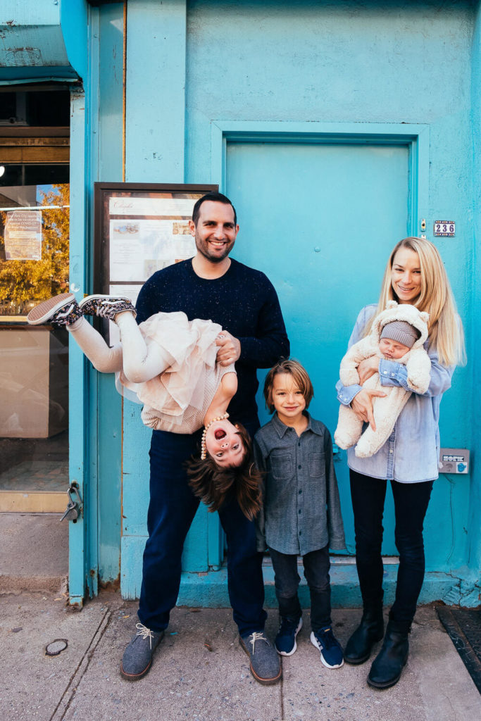 A family of five joyfully poses in front of a turquoise wall. The vibrant scene, captured by a talented Brooklyn family photographer, shows a man playfully holding a girl upside down, while a woman cradles a baby and a boy smiles between them.