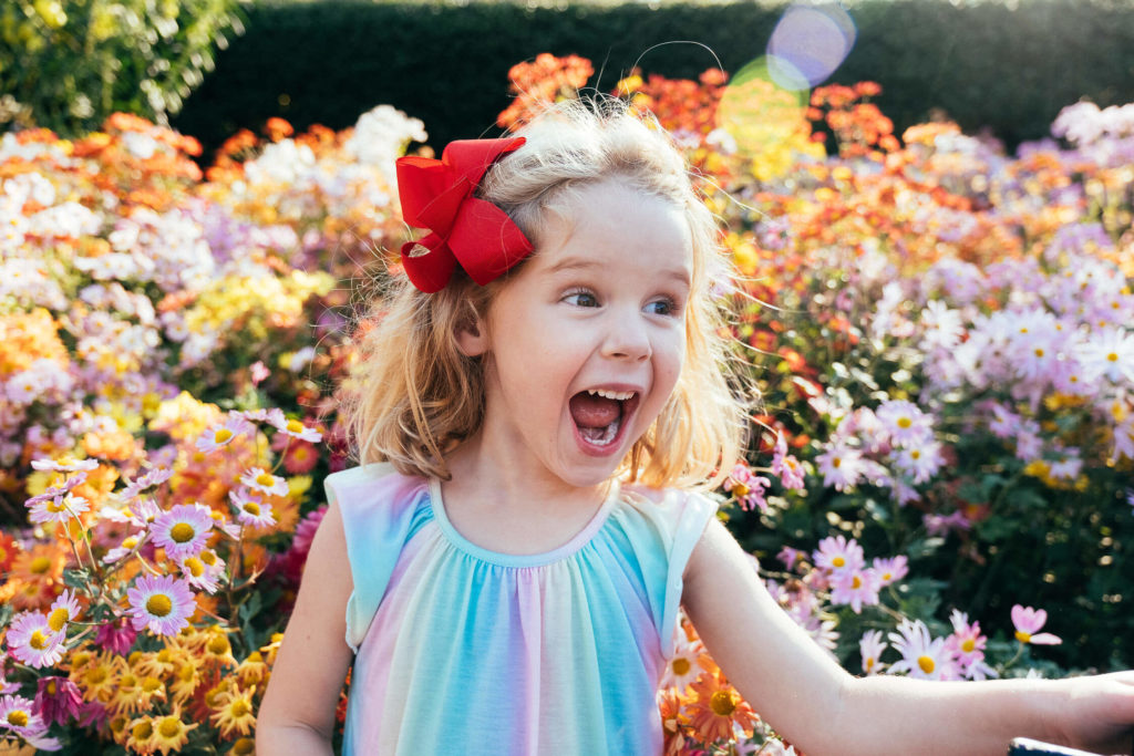 smiling girl with a red bow in her hair, surrounded by vibrant flowers in a garden setting