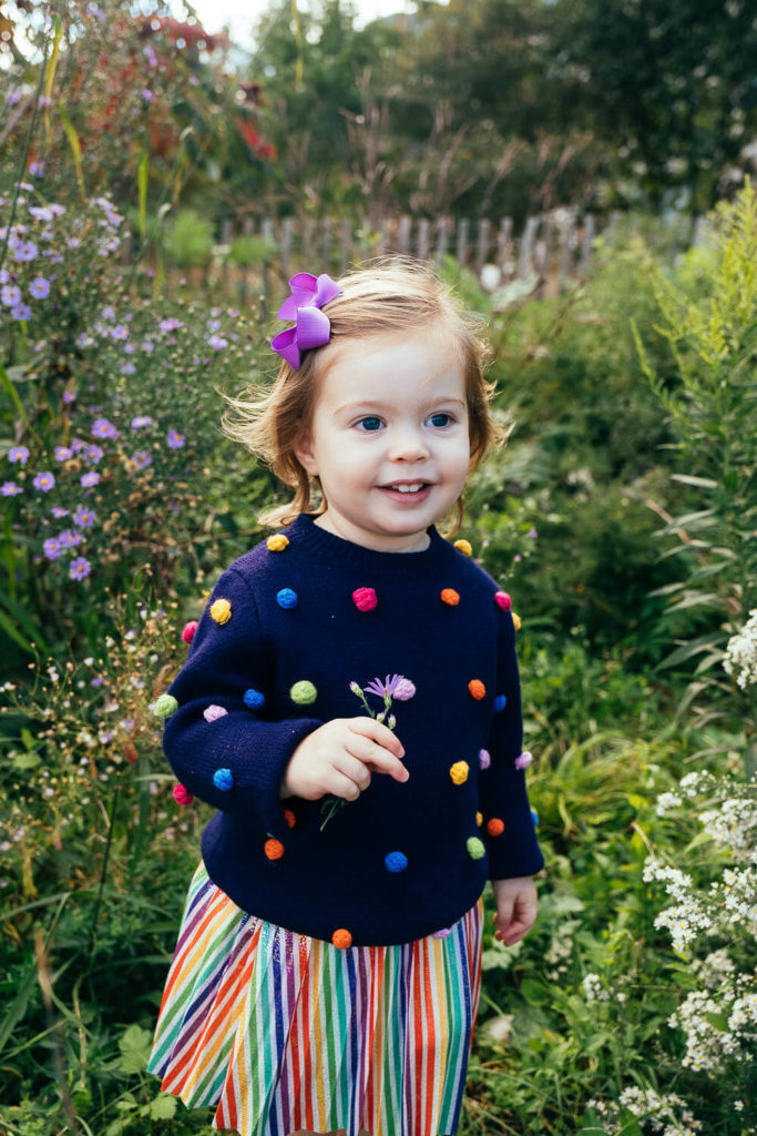 Little girl with a purple flower in her hair and a colorful polka dot sweater, standing in a garden.