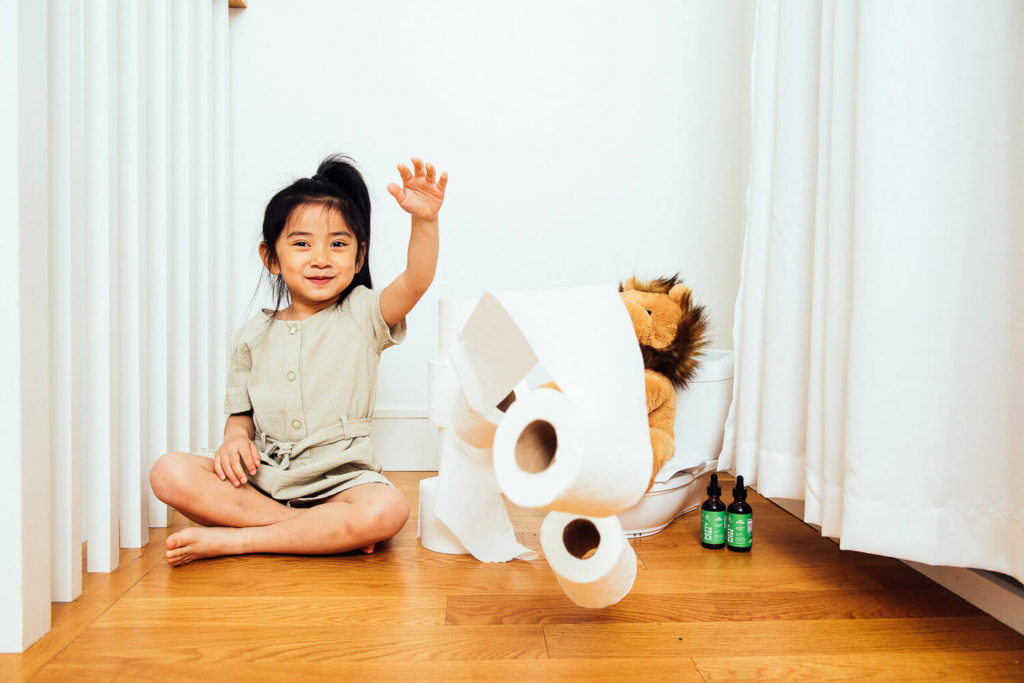 A child sits on the floor, waving cheerfully. Next to her, a stuffed animal perches on a toilet seat surrounded by rolls of toilet paper, with bottles scattered nearby. The scene exudes light-hearted charm and could be straight out of a playful brand photography session.