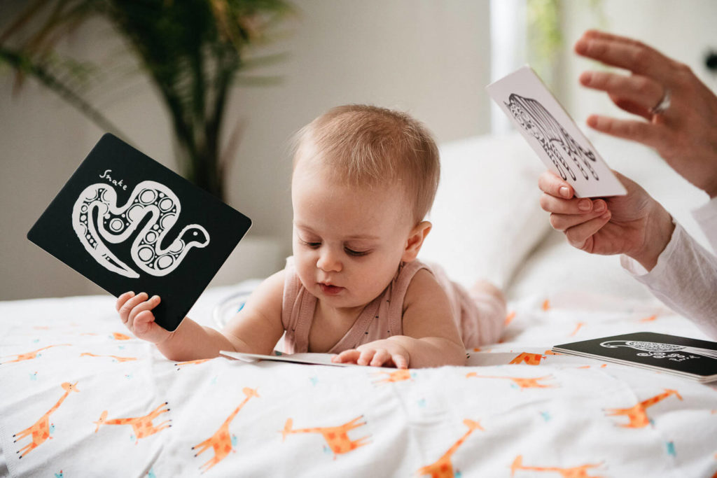 A baby rests on a bed with giraffe-patterned sheets, holding a card featuring a black and white snake illustration—an example of brand photography. Another hand displays a similar card depicting a hand, while a plant adds natural charm to this commercial photography setup.