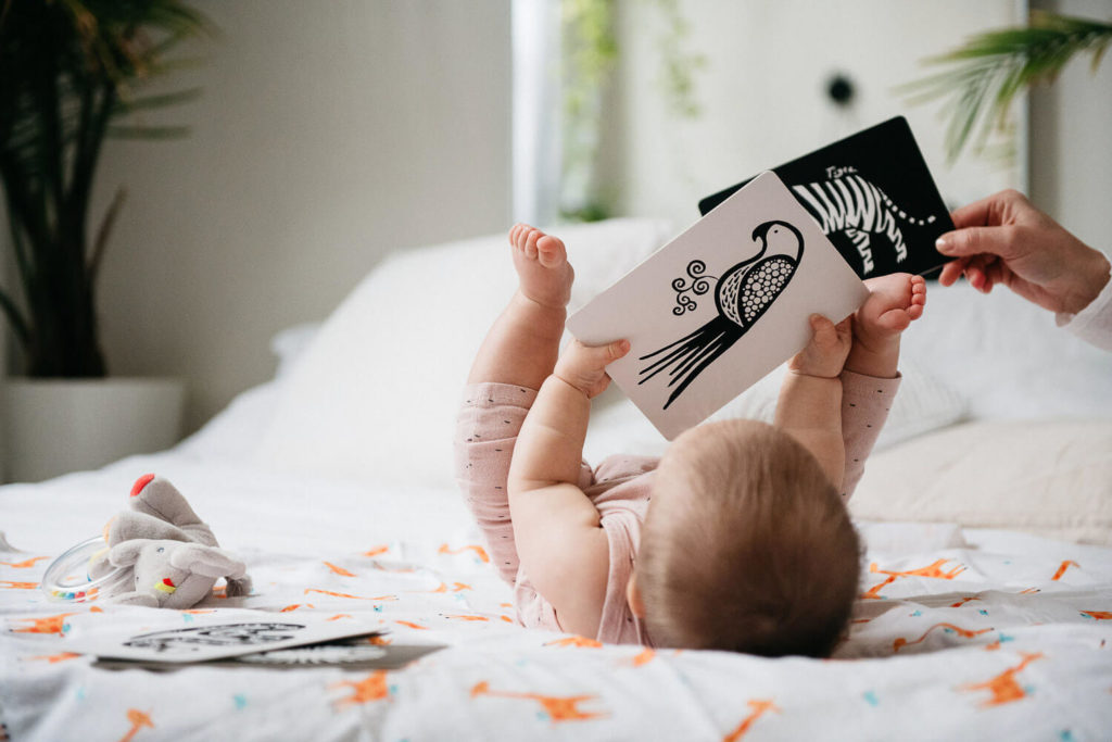 A baby lying on a bed holds up a black and white card featuring an illustration of a bird, showcasing the essence of brand photography. Other animal illustration cards are scattered around, enhancing the room's bright, airy feel with plants in the background.