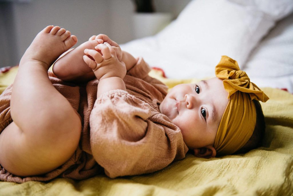A baby in a mustard yellow headband lies on a yellow blanket, holding its feet in delight. Dressed in a peach-colored outfit, the content infant is framed perfectly for brand photography. White pillows softly accentuate the background.