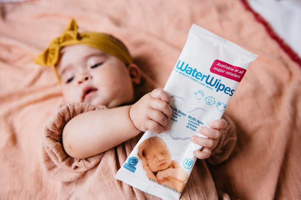 A baby wearing a yellow headband lies on a peach blanket, holding a pack of WaterWipes. Capturing the essence of brand photography, the wipes package displays an image of a sleeping baby and text highlighting 99.9% water content.