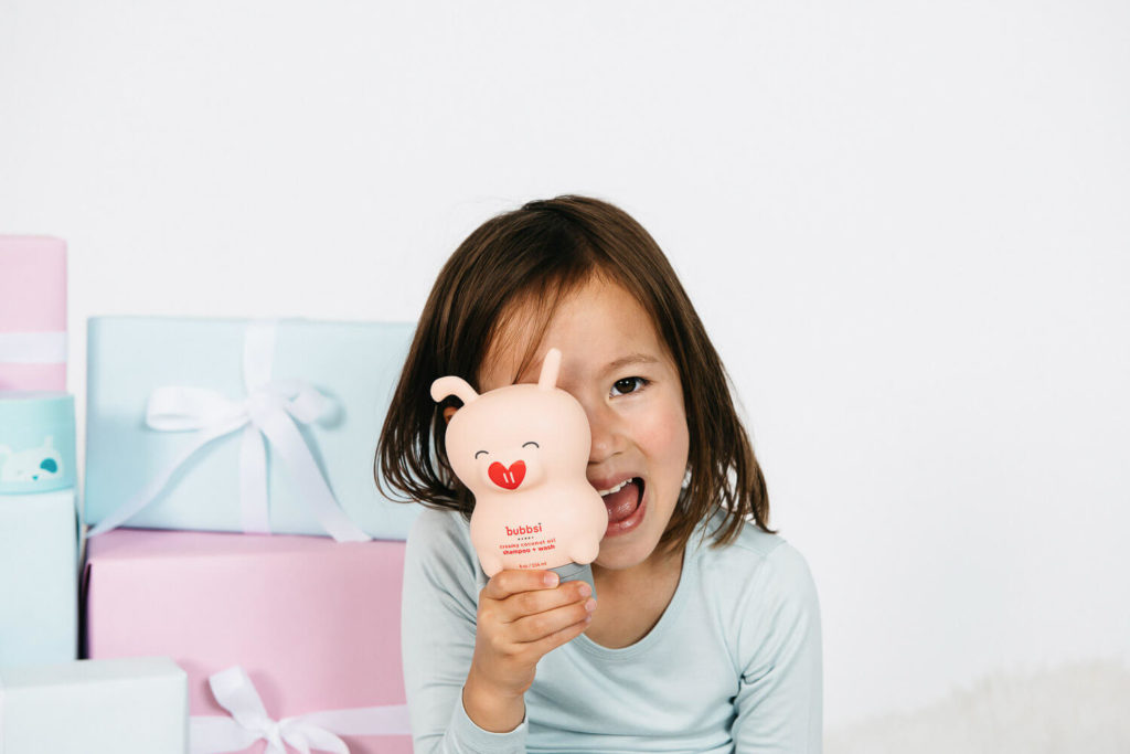 A young child with brown hair joyfully smiles while clutching a cute beige toy adorned with a red heart-shaped nose. The child, dressed in a light blue shirt, stands amidst an array of pastel-colored gift boxes with ribbons—an enchanting scene perfect for brand photography.