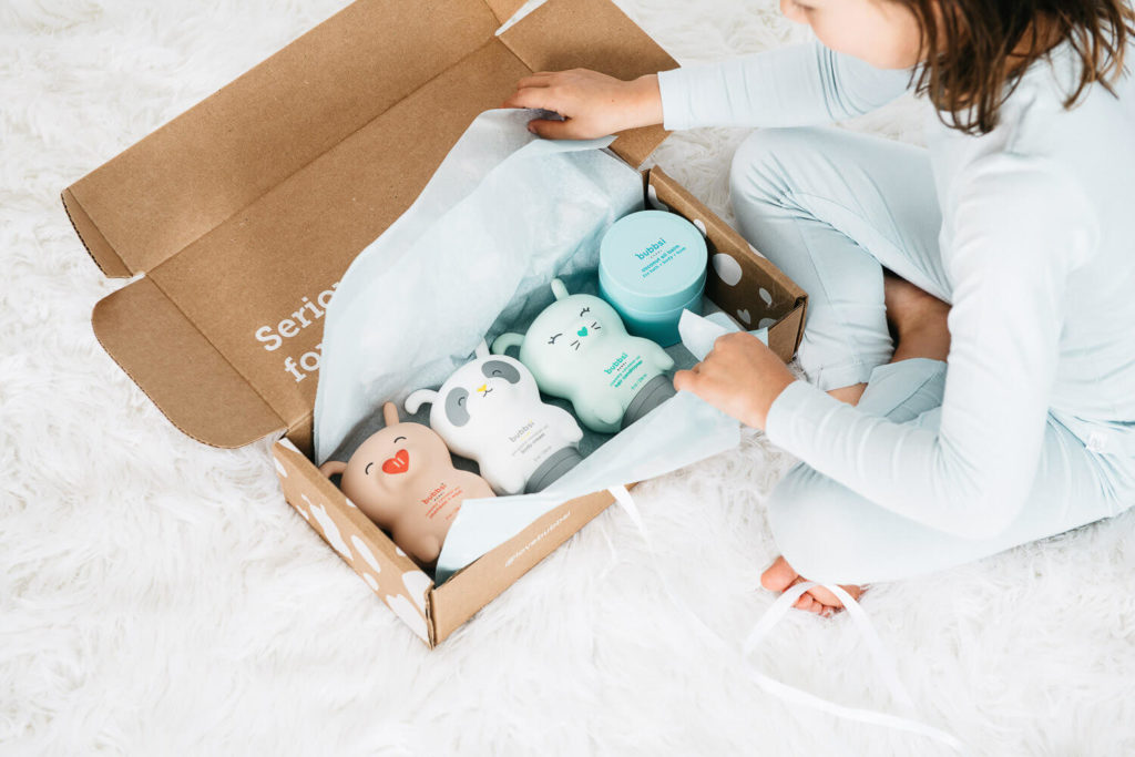 A child in light blue pajamas sits on a fluffy white rug, unboxing a cardboard package with four pastel-colored, animal-themed bottles. This delightful scene captures the essence of brand photography, showcasing bottles with playful, smiling faces nestled in tissue paper.