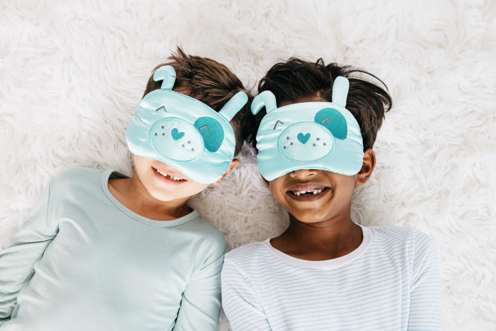 Two children lying on a fluffy white rug, smiling and wearing matching blue animal-themed sleep masks with cute designs and ear patterns capture the essence of brand photography.