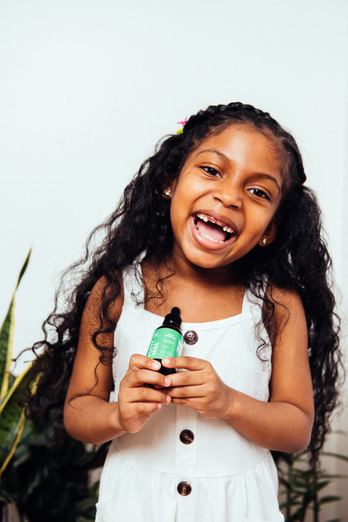 A young girl with long curly hair smiles widely, holding a green bottle in what could easily be a brand photography session. She wears a white dress with brown buttons, and in the background, lush green leaves add to the commercial photography appeal of the scene.