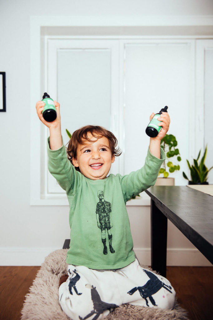A cheerful child with light brown hair sits on a chair, showcasing their joy in brand photography. They're wearing a green shirt and patterned pants, holding two small bottles in each hand with a radiant expression. The white wall and window with partially open blinds complete the scene.