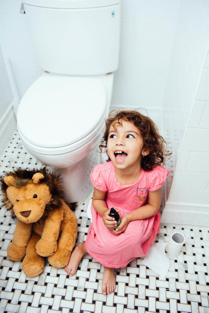 A little girl in a pink dress sits on the bathroom floor next to a toilet, holding a small object. A plush lion toy is beside her, and a roll of toilet paper is on the floor. Capturing her joyful expression, this scene embodies the charm of brand photography.