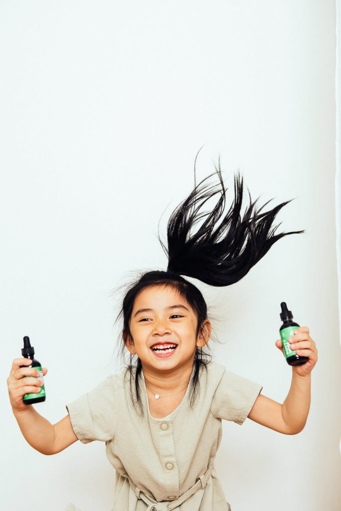 A joyful child with long black hair jumps playfully, holding two small bottles with dropper caps in her hands. Clad in a light-colored dress and flashing a big smile against a plain background, she embodies the essence of brand photography.