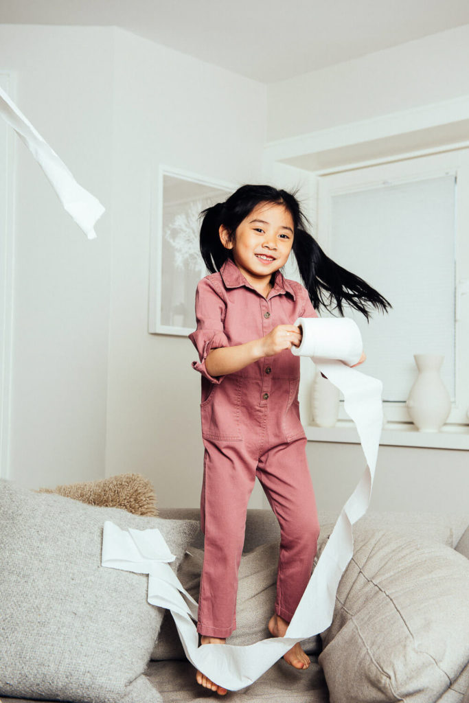 A young girl with black hair, wearing a pink jumpsuit, joyfully jumps on a couch while unrolling a roll of toilet paper—a playful moment captured perfectly for brand photography. The room features neutral tones, a window with white blinds, and a framed picture on the wall.