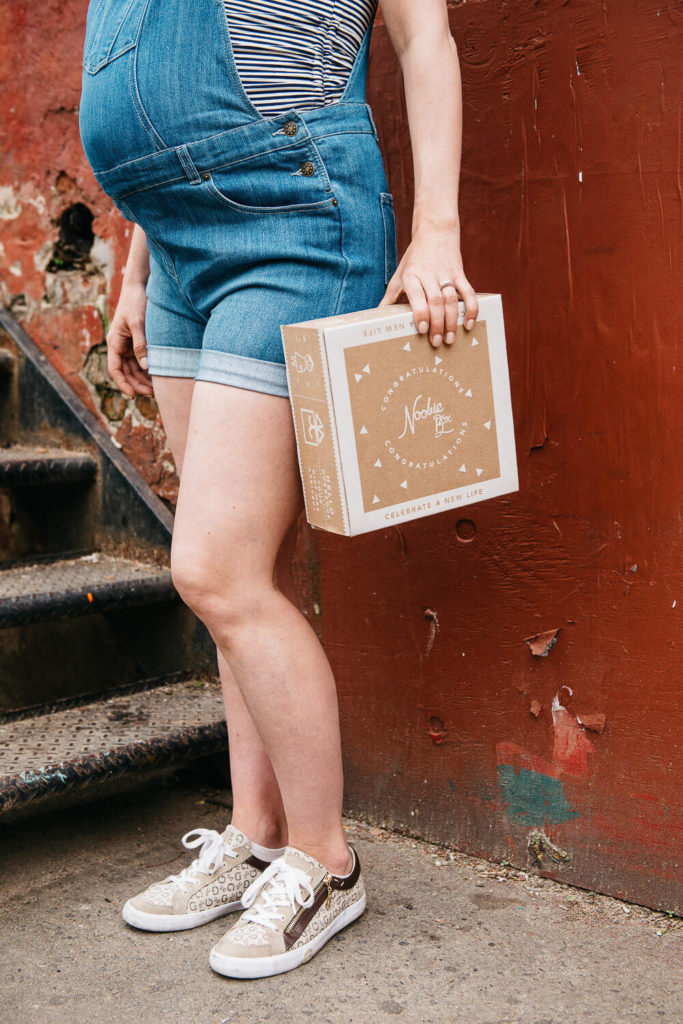 A pregnant person in denim overalls stands beside a rusted staircase, holding a Noobie Box adorned with baby illustrations, embodying a casual yet thoughtful brand photography scene. Their sneakers and striped shirt add charm against the worn red wall backdrop.