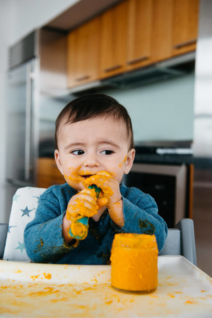 A baby sits in a high chair, joyfully eating with hands, covered in orange food. An open jar of food rests on the tray. The kitchen, perfect for commercial photography, showcases modern wooden cabinets and stainless steel appliances in the background.
