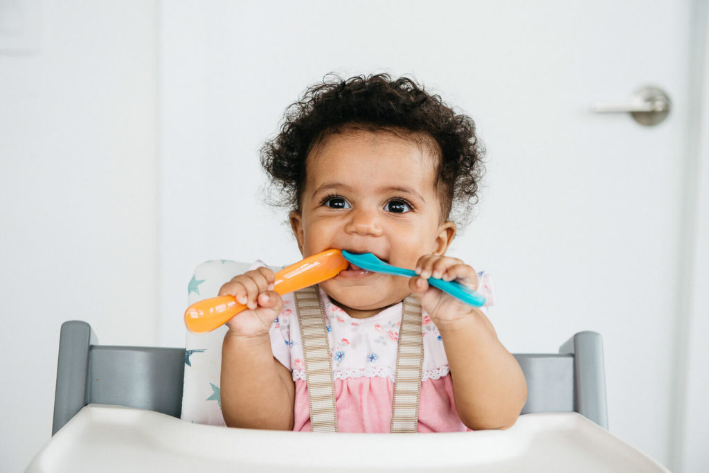 A cheerful baby with curly hair sits in a high chair, wearing a pink outfit suitable for brand photography. The little one holds an orange and a blue spoon, smiling at the camera—a perfect moment captured in commercial photography. A light-colored wall with a closed door forms the background.