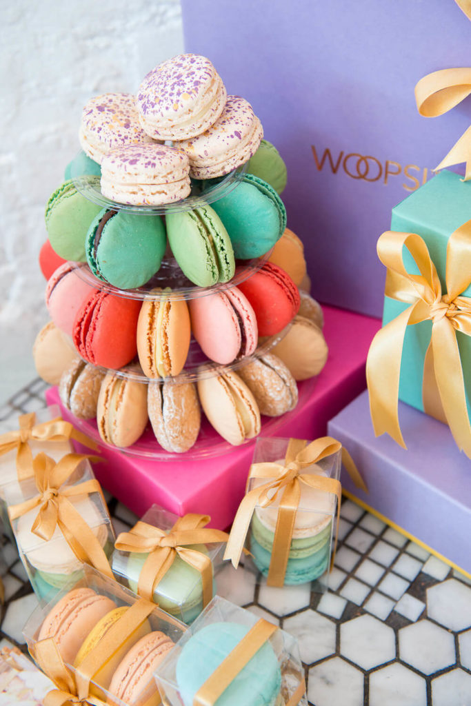 A colorful tower of assorted macarons on a glossy stand is surrounded by elegantly wrapped gift boxes. Perfect for brand photography, these pastel macarons in purple, green, pink, and beige are beautifully packed in small clear boxes tied with golden ribbons.