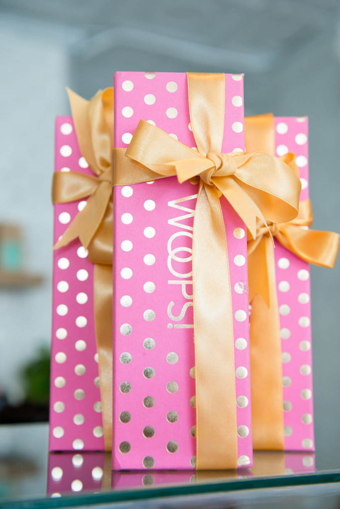 Three pink gift boxes with gold polka dots, wrapped in gold satin bows, are elegantly stacked on a glass surface—a perfect display of brand photography.