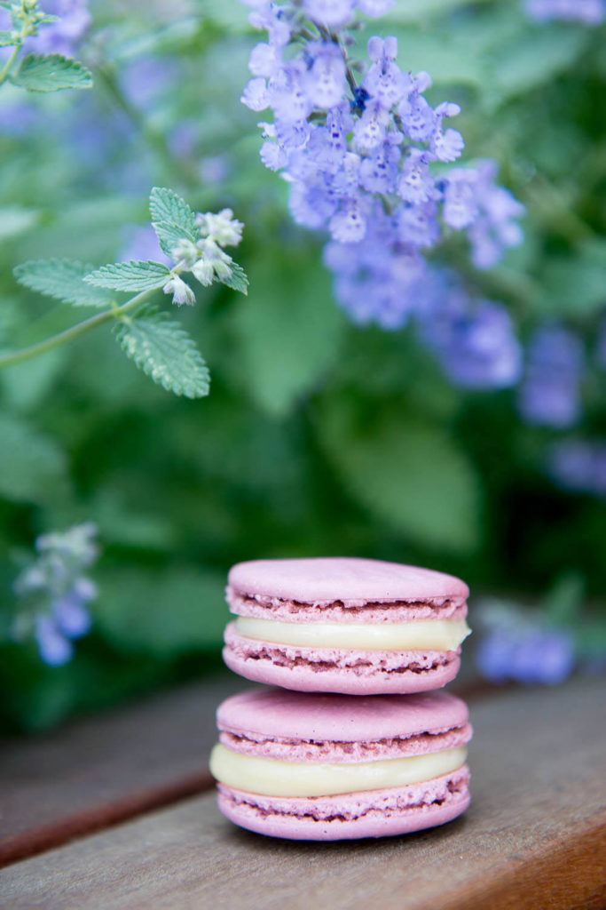 Two pink macarons stacked on a wooden surface capture the elegance of brand photography, with a blurred background of green leaves and small purple flowers that adds a touch of natural beauty.