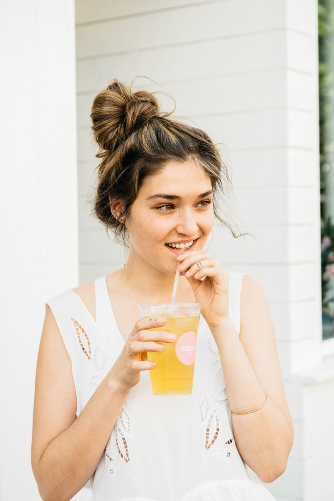 A woman with a messy bun sips a cold drink through a straw, showcasing her casual style. In a white sleeveless top against a stark white wall, she embodies effortless charm—an ideal subject for brand photography—smiling slightly as she gazes off to the side.