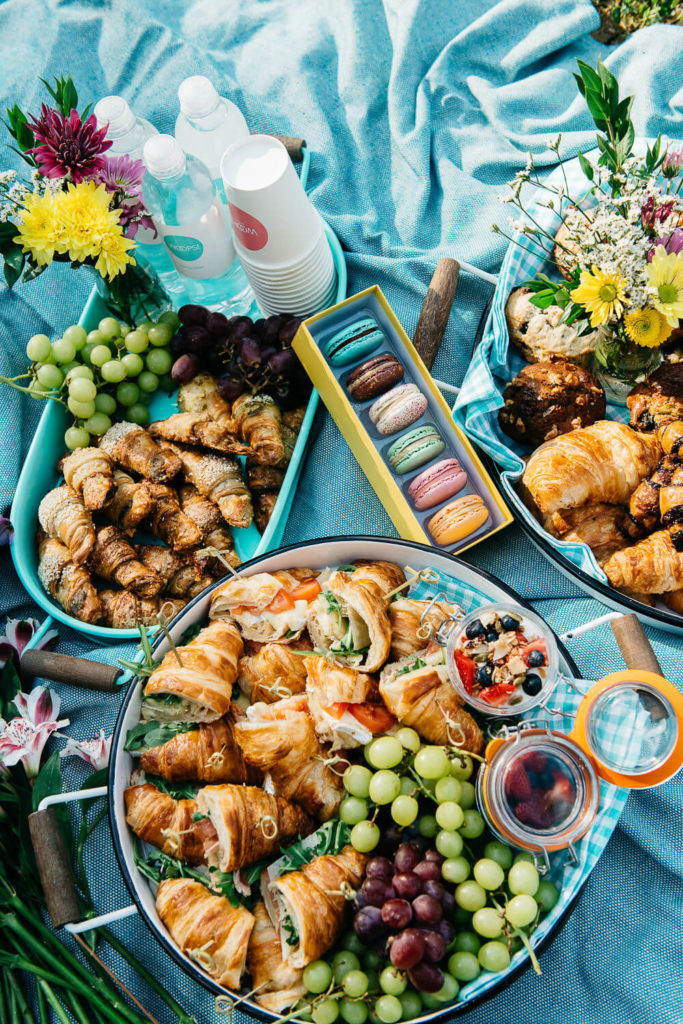 A vibrant picnic spread on a blue blanket showcases trays of croissants, macarons, grapes, and bottled water. Fresh flowers and pastries add colorful touches to this perfect moment for brand photography.