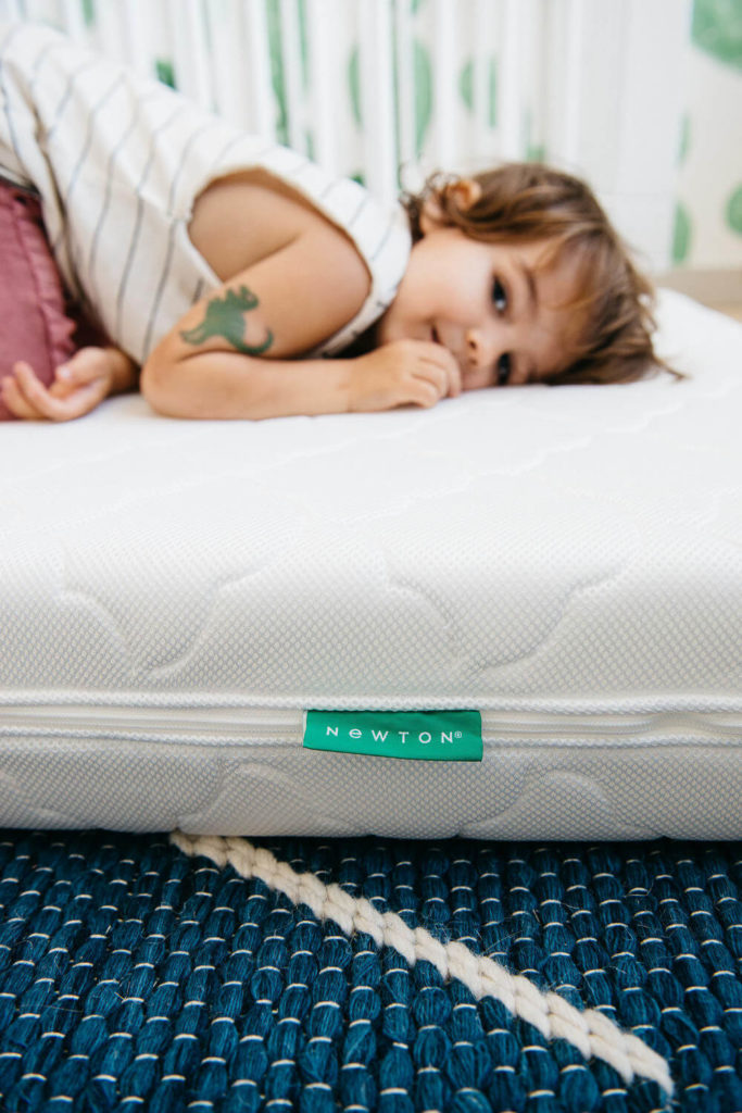 A child lies on a white mattress with a green Newton label, perfect for brand photography. The child, sporting short brown hair, wears a striped shirt and shows off a dinosaur tattoo on their arm. The scene sits atop a blue woven rug, capturing commercial photography at its finest.