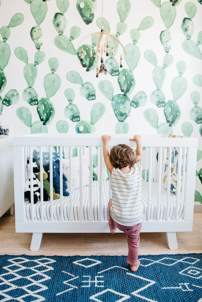 A toddler in a striped shirt and pink pants climbs into a white crib in a nursery, perfect for commercial photography. The wall boasts a green cactus pattern, with a mobile hanging above the crib. The floor features a dark blue patterned rug, tying the room's aesthetic together beautifully.