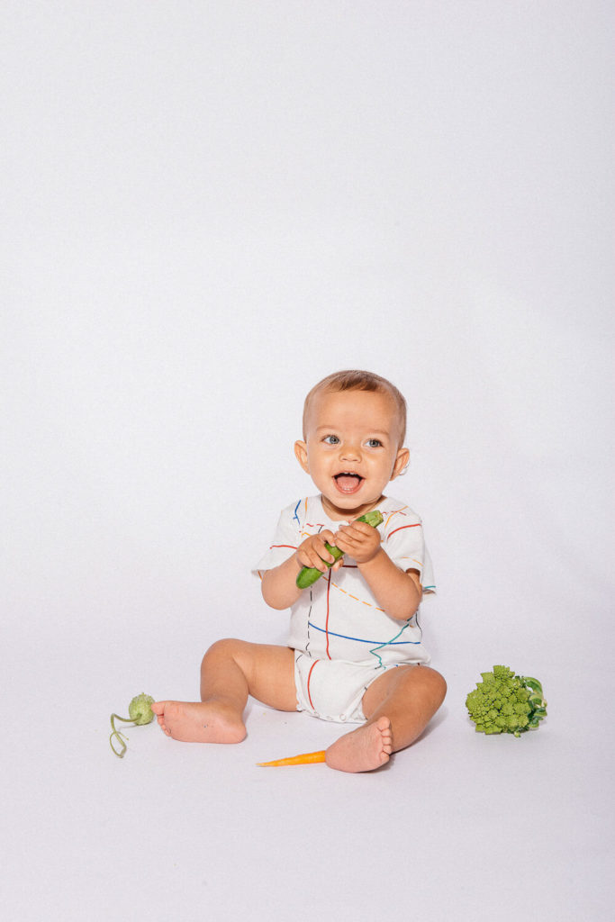 A happy baby sits on the floor in a white outfit with colorful lines, holding a piece of broccoli. Perfect for brand photography, a broccoli head and a carrot lie beside the baby against a plain white backdrop, capturing an authentic moment ideal for commercial photography.