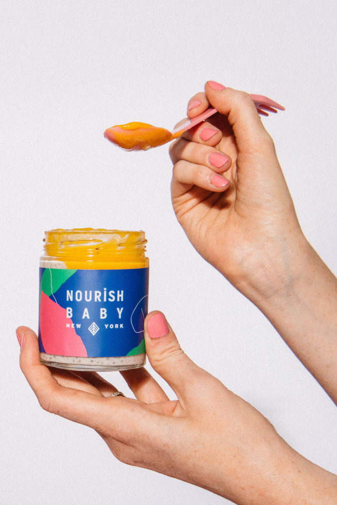 A person with pink nail polish holds a spoonful of orange baby food from a jar labeled Nourish Baby New York, showcasing vibrant brand photography against a plain white background.