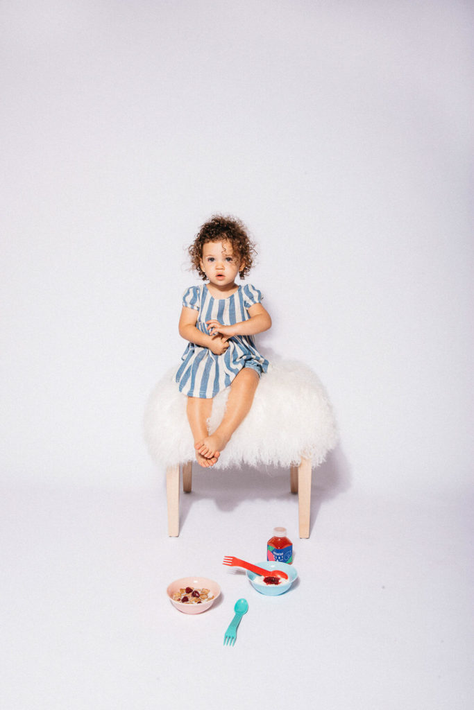A young child with curly hair sits on a small, fluffy stool wearing a blue and white striped dress. In this slice of brand photography, the scene features a bowl of cereal, a cup, and utensils on the floor against a plain background.