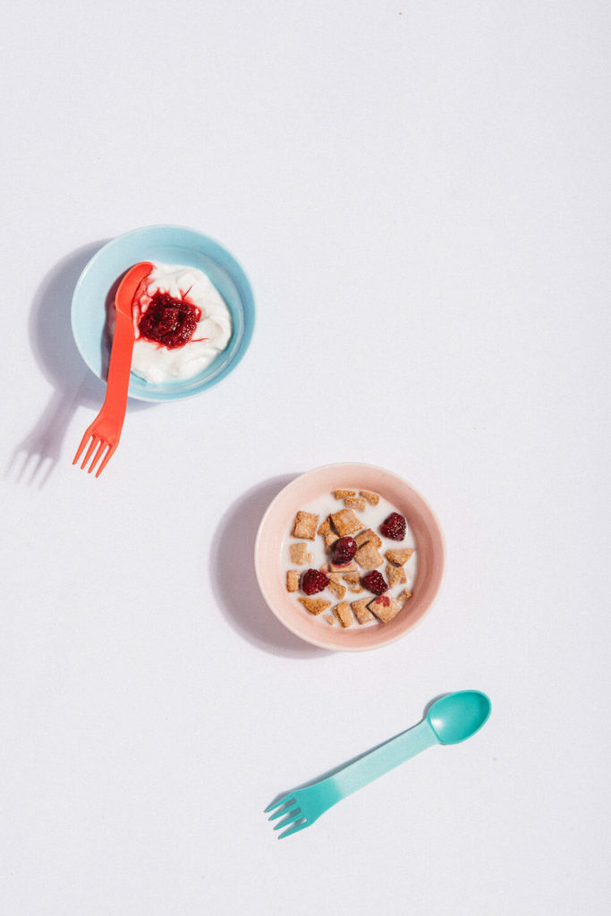 Two bowls set on a white surface, each containing yogurt, perfect for brand photography. One is topped with fruit preserves and a red fork, while the other has cereal pieces and red berries, with a blue spoon. A minimalist, overhead view ideal for commercial photography.