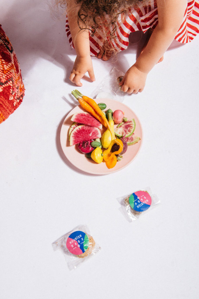 A child in a red and white striped outfit is sitting on a white surface, carefully arranging a colorful plate of vegetables, like carrots, radishes, and squash. This moment of artistry shines through brand photography as two small packaged snacks lie nearby.