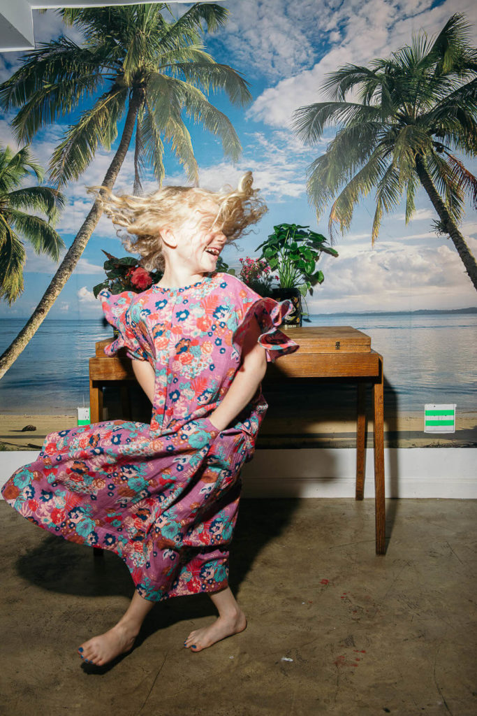 A barefoot child with blonde hair in motion, wearing a colorful floral dress, dances joyfully in front of a tropical beach backdrop with palm trees and a blue sky. Perfect for brand photography, the scene captures vibrant energy against a wooden table adorned with plants and flowers.