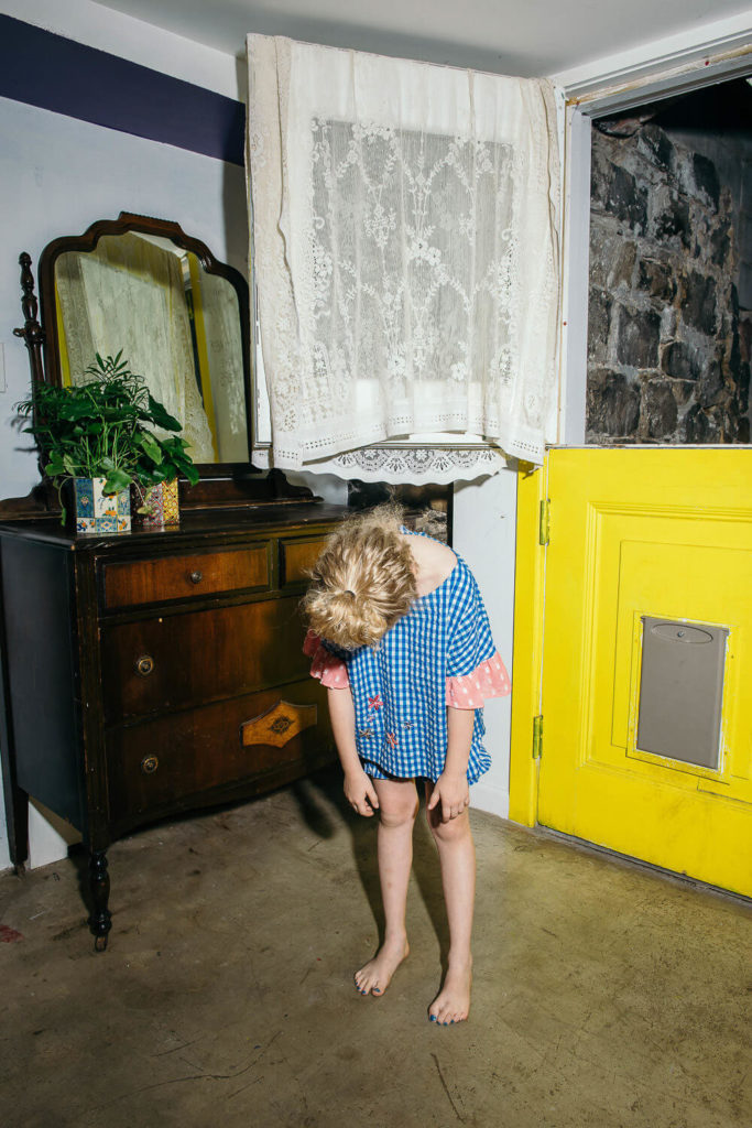 A child in a blue checkered dress with pink polka-dot sleeves stands barefoot, bending forward with their head down. Set in a room reminiscent of brand photography, there's a wooden dresser, mirror, plant, and yellow door adorned with lace curtains above.