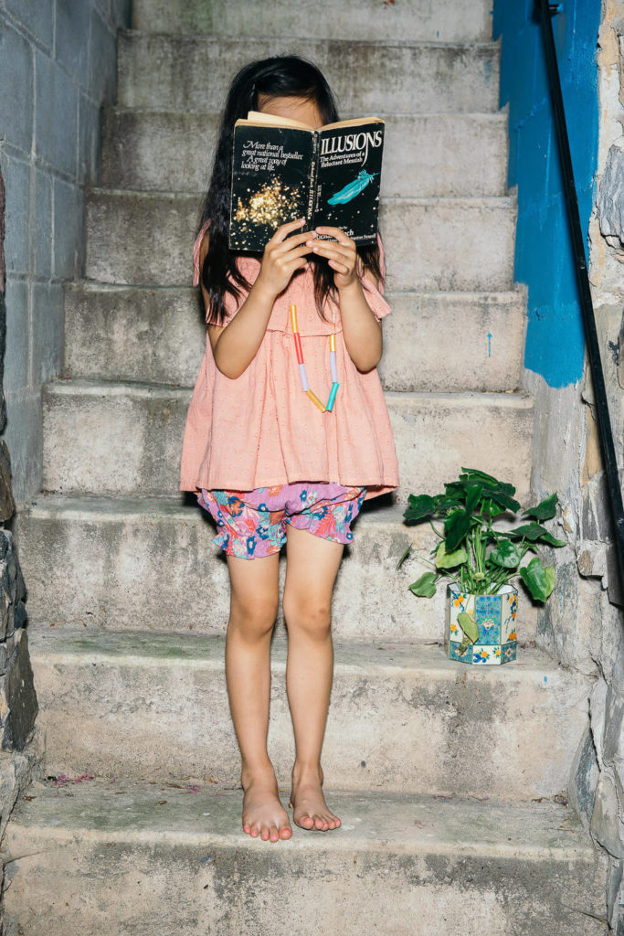 A barefoot girl poses on concrete stairs for a brand photography shoot, concealing her face with a book titled 
