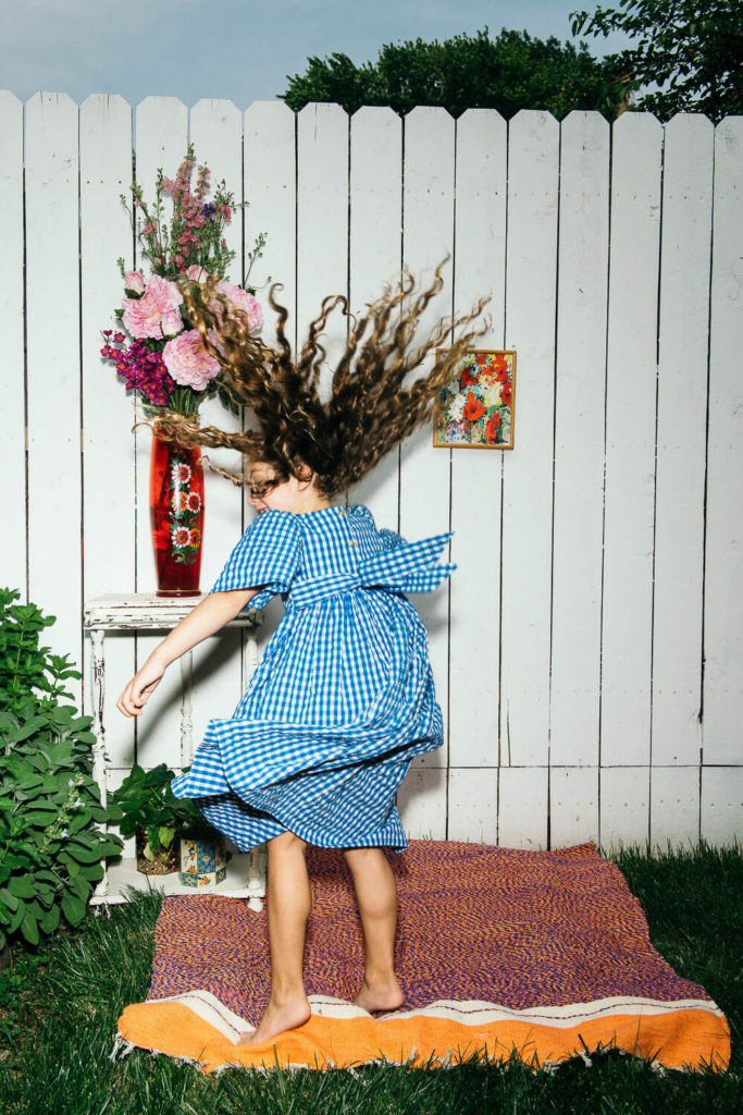 A child in a blue checkered dress joyfully jumps, her curly hair dancing against the backdrop of a white picket fence. This charming scene captures the essence of brand photography, with an orange rug underfoot and artful details like a flower vase enhancing the vibrant greenery.