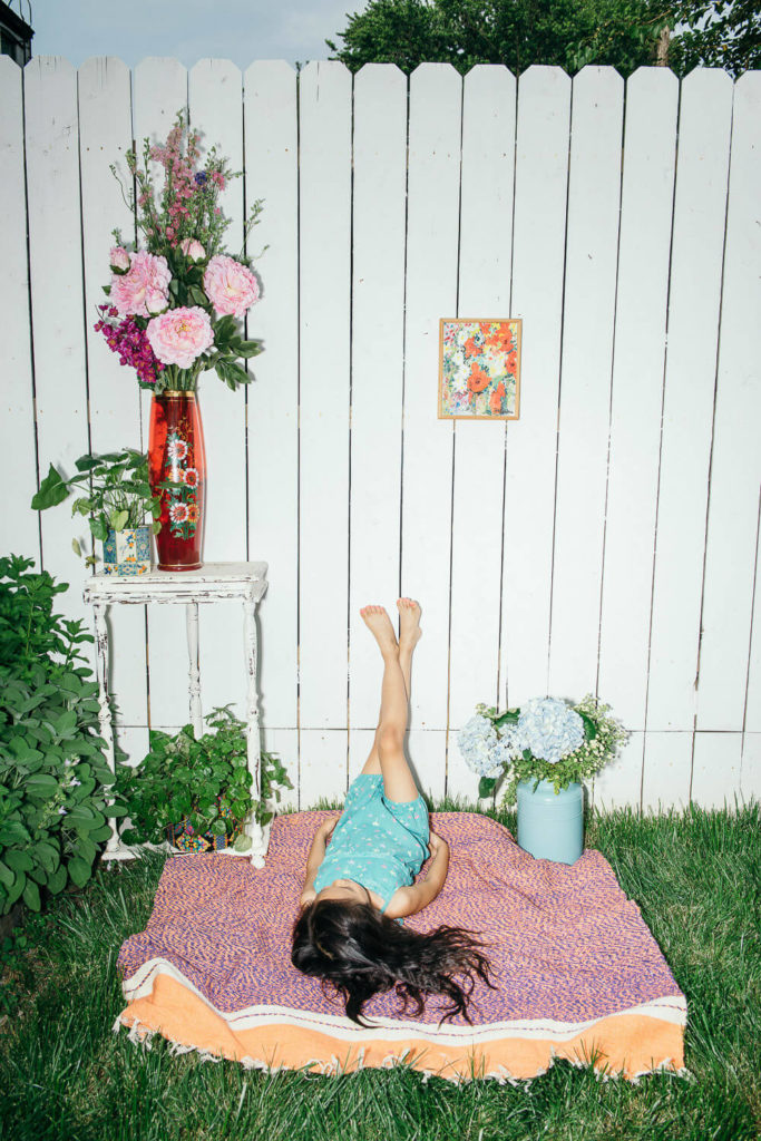 A child lies on a blanket in a backyard, legs up against a white picket fence. Nearby are flowers in a vase on a table and a teal bucket. A colorful picture hangs on the fence, capturing the essence of brand photography with its bright and summery scene.