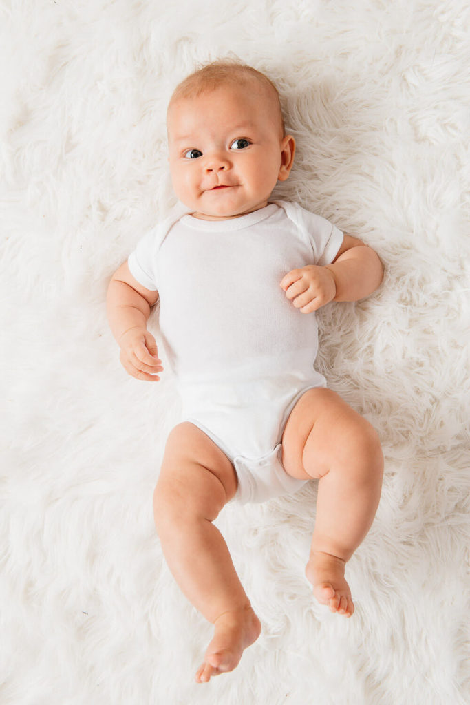 A baby in a white onesie lies on a fluffy white rug, looking up with a gentle smile. With short hair and chubby cheeks, the child exudes warmth and innocence—ideal for brand photography that captures pure, heartfelt moments.