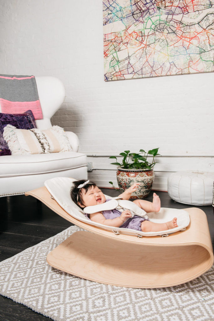 A baby in a purple outfit is happily lounging in a modern wooden rocker on a patterned rug. Captured through commercial photography, the room showcases a white armchair with colorful cushions, a potted plant, and an abstract map artwork on the wall.