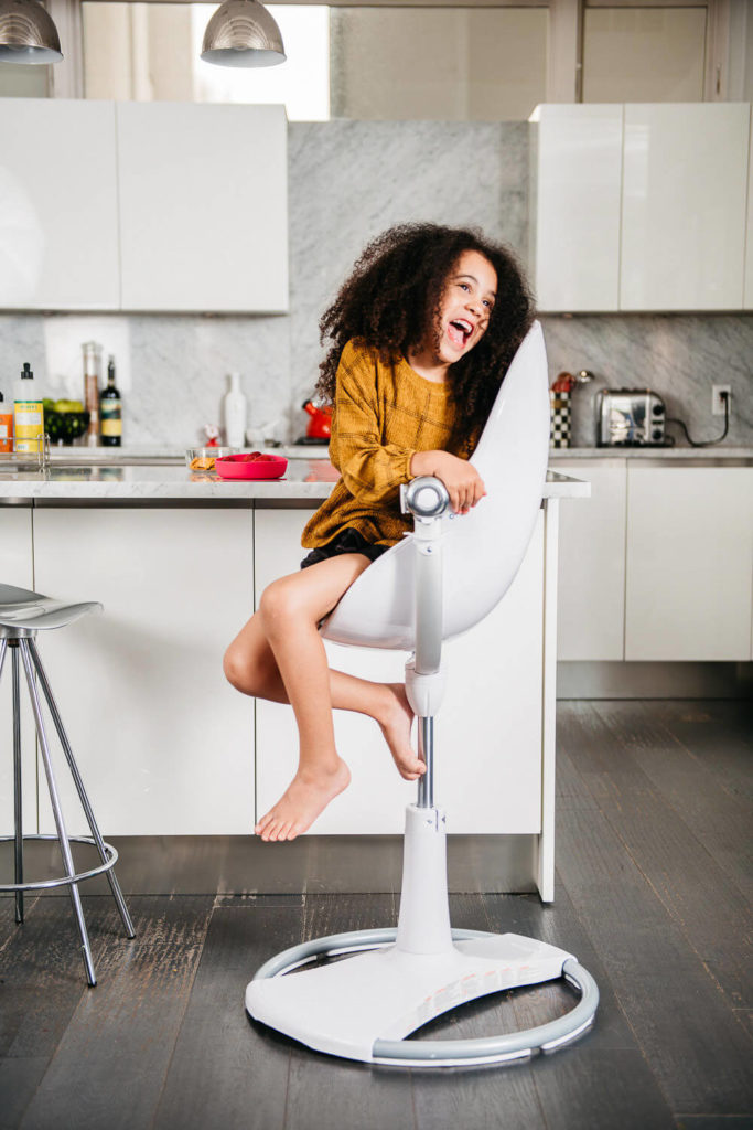 A young girl with curly hair and a brown dress is laughing while sitting on a modern white swivel chair in a bright kitchen, embodying the essence of brand photography. She has one leg bent and is leaning back, enjoying herself amid the white cabinets and marble countertop.
