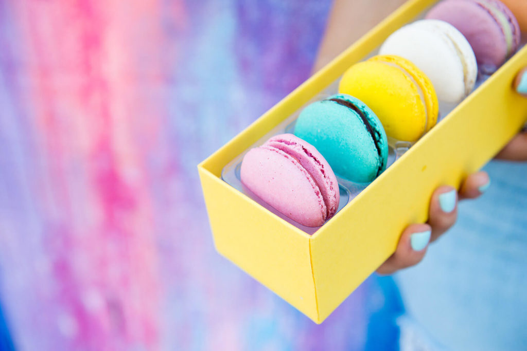 In a vibrant display of brand photography, a person with light blue painted nails holds a yellow box containing five colorful macarons against a lively pink and blue backdrop. The neatly arranged macarons—pink, blue, yellow, white, and another blue—add to the visual feast.
