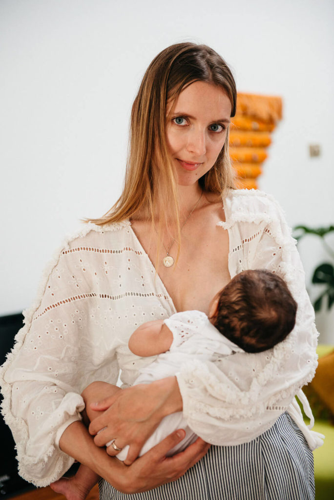 A woman with long hair, wearing a white blouse, lovingly holds a baby in her arms in this beautiful example of brand photography. They are indoors, highlighted against a blurred background featuring a plant and an orange accent piece.